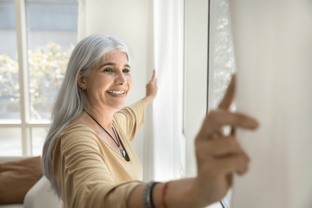 Inspired mature grey-haired woman standing near window and opening curtains, looking outside with smile.