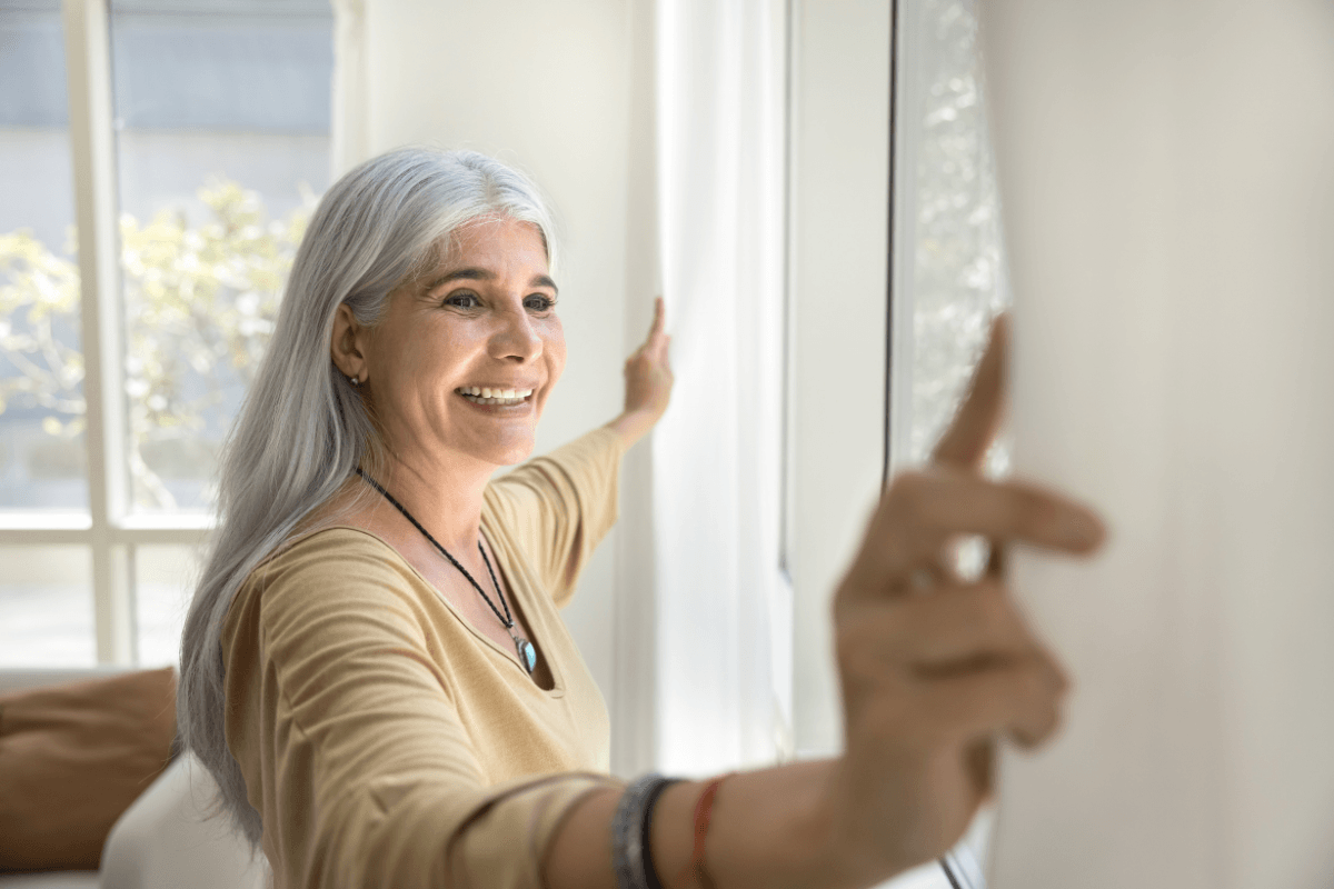 Inspired mature grey-haired woman standing near window and opening curtains, looking outside with smile.